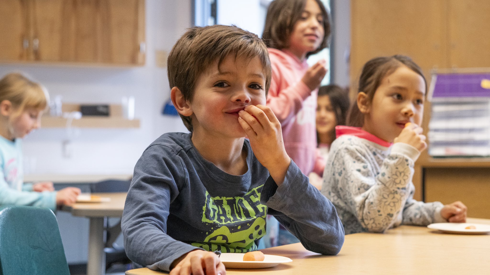 An image of a child eating some food. Two children are seated at this table and the boy is the focus of this image and is quite happy. This image is used as our Feeding Futures Header image